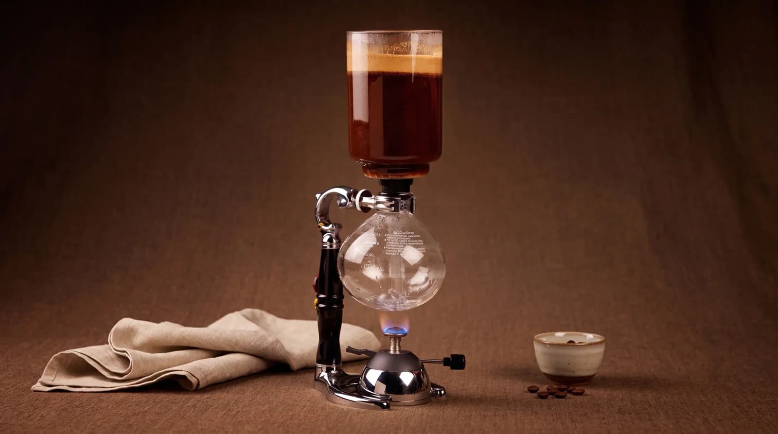 Halogen glow under a siphon's lower globe, brew rising like a lava lamp.